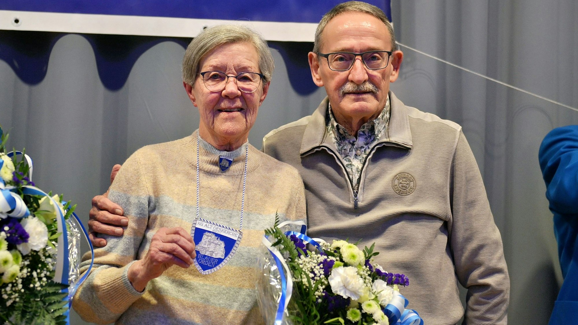 Ordensträgerin Ursula Kothe zeigt den Blauen Orden, den sie um den Hals trägt. Ihr Lebensgefährte steht neben ihr und hält sie im Arm.