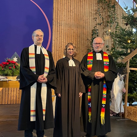 Zwei Männer und eine Frau stehen im schwarzen Ornat im Altarraum einer Kirche.