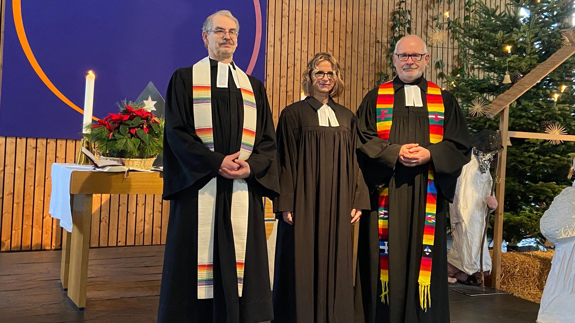 Zwei Männer und eine Frau stehen im schwarzen Ornat im Altarraum einer Kirche.