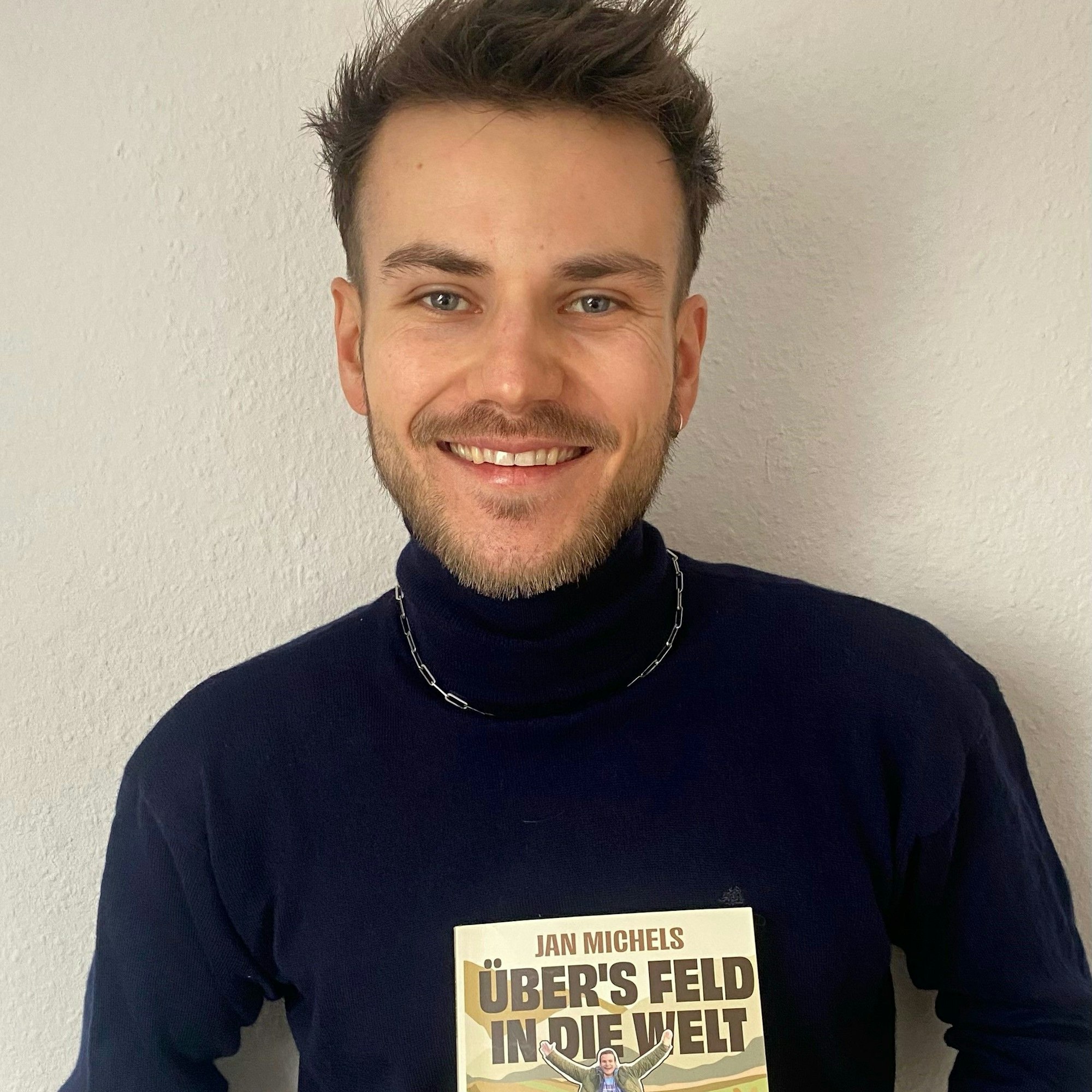 Jan Michels mit seinem Buch „Übers Feld in die Welt“. Er trägt einen dunkelblauen Rollkragenpullover mit silberner Kette.