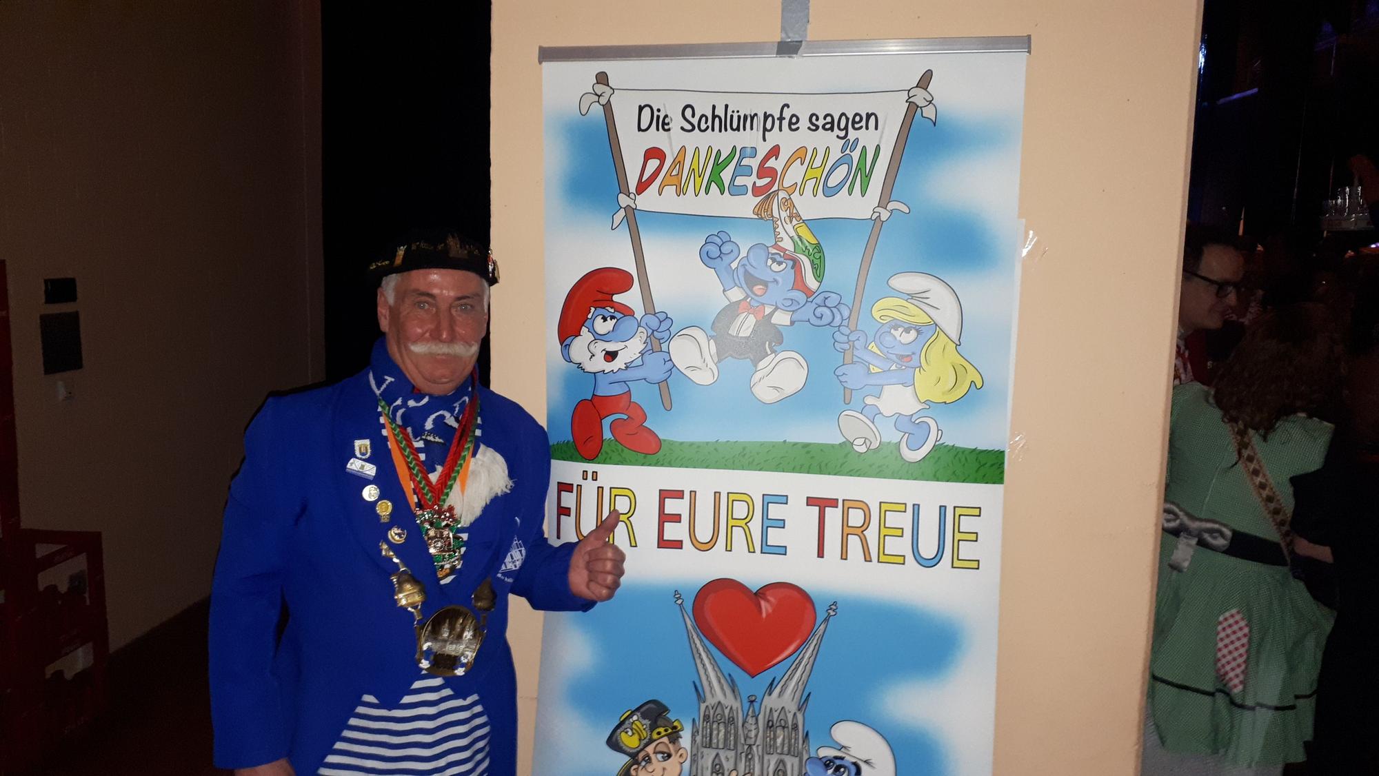 Ein Mann mit einem Schnauzer steht vor einem Plakat, auf dem Schlümpfe drauf sind und steht, Die Schlümpfe sagen Dankeschön für eure Treue.