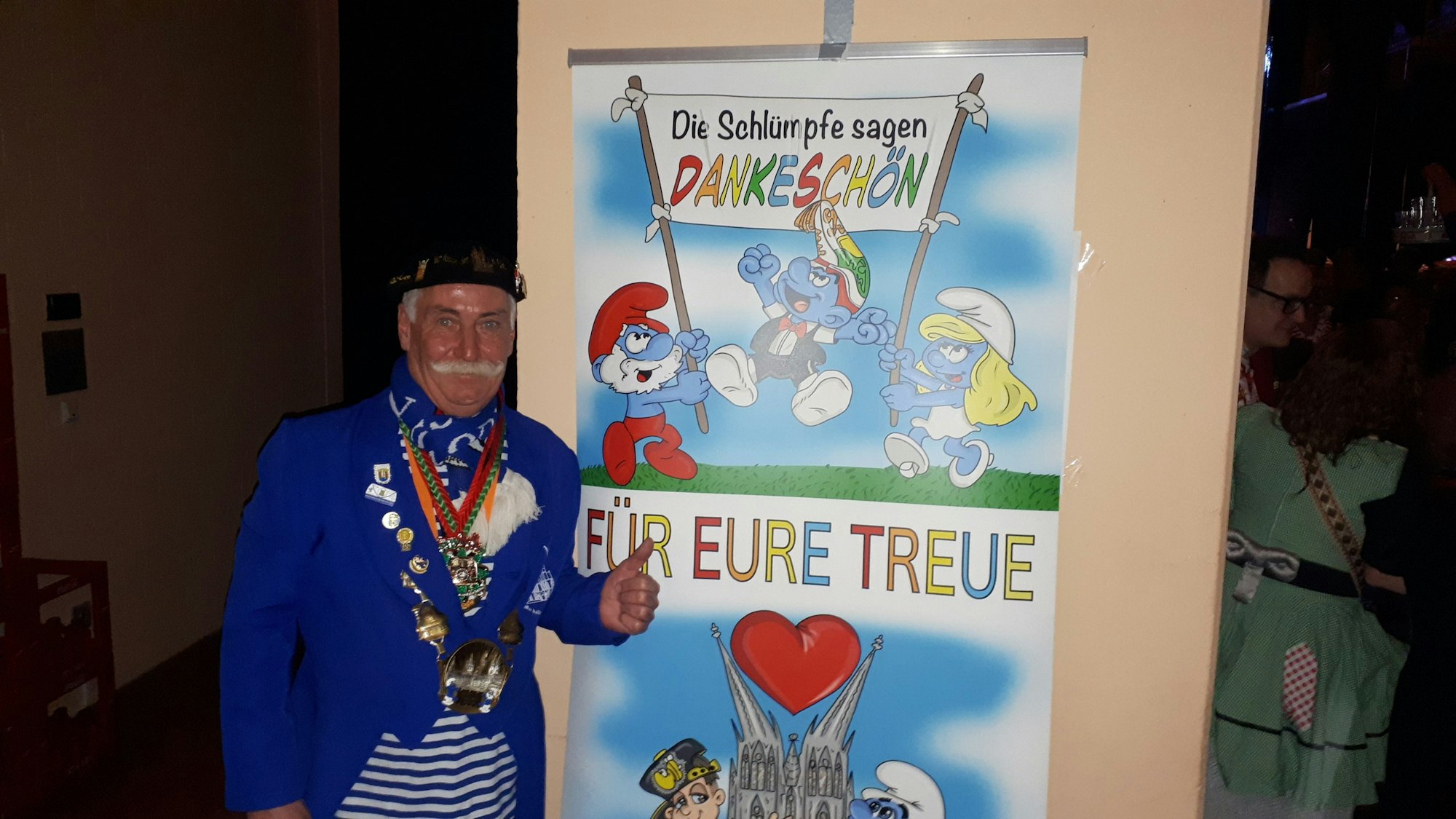 Ein Mann mit einem Schnauzer steht vor einem Plakat, auf dem Schlümpfe drauf sind und steht, Die Schlümpfe sagen Dankeschön für eure Treue.