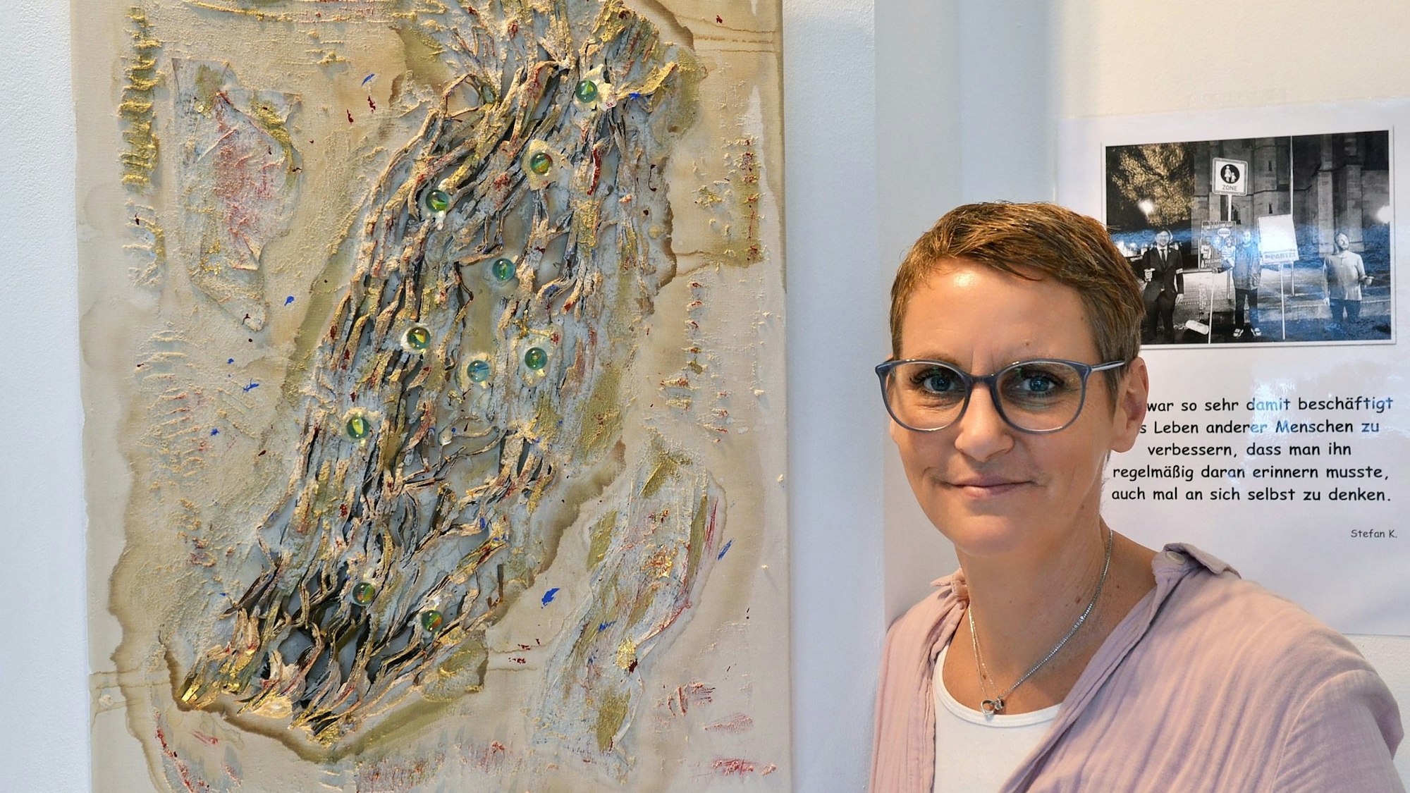 Nicole Dreifke steht neben einem Werk des Künstlers, das an einer Wand hängt.