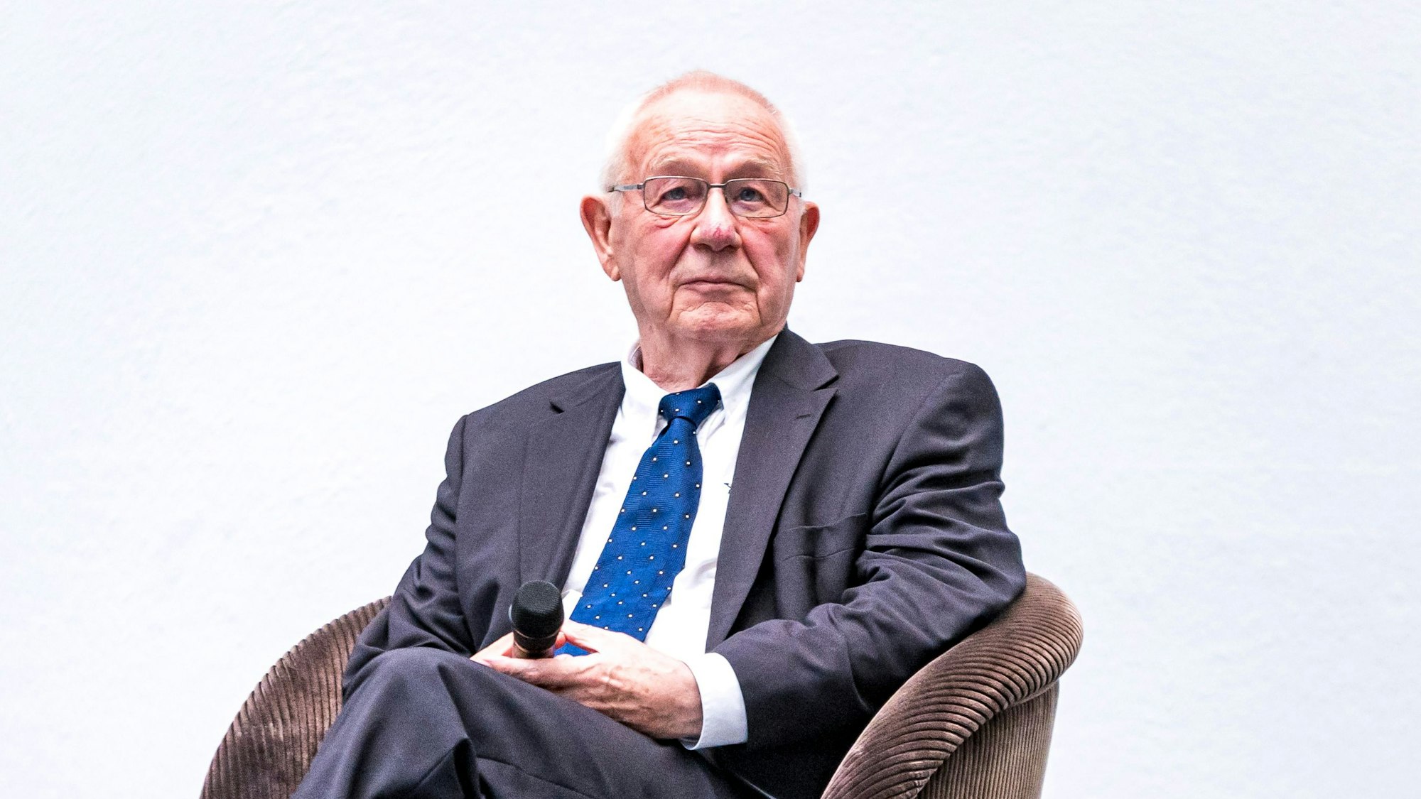 Armin Maiwald sitzt im Anzug und mit Mikrofon auf einer Bühne.