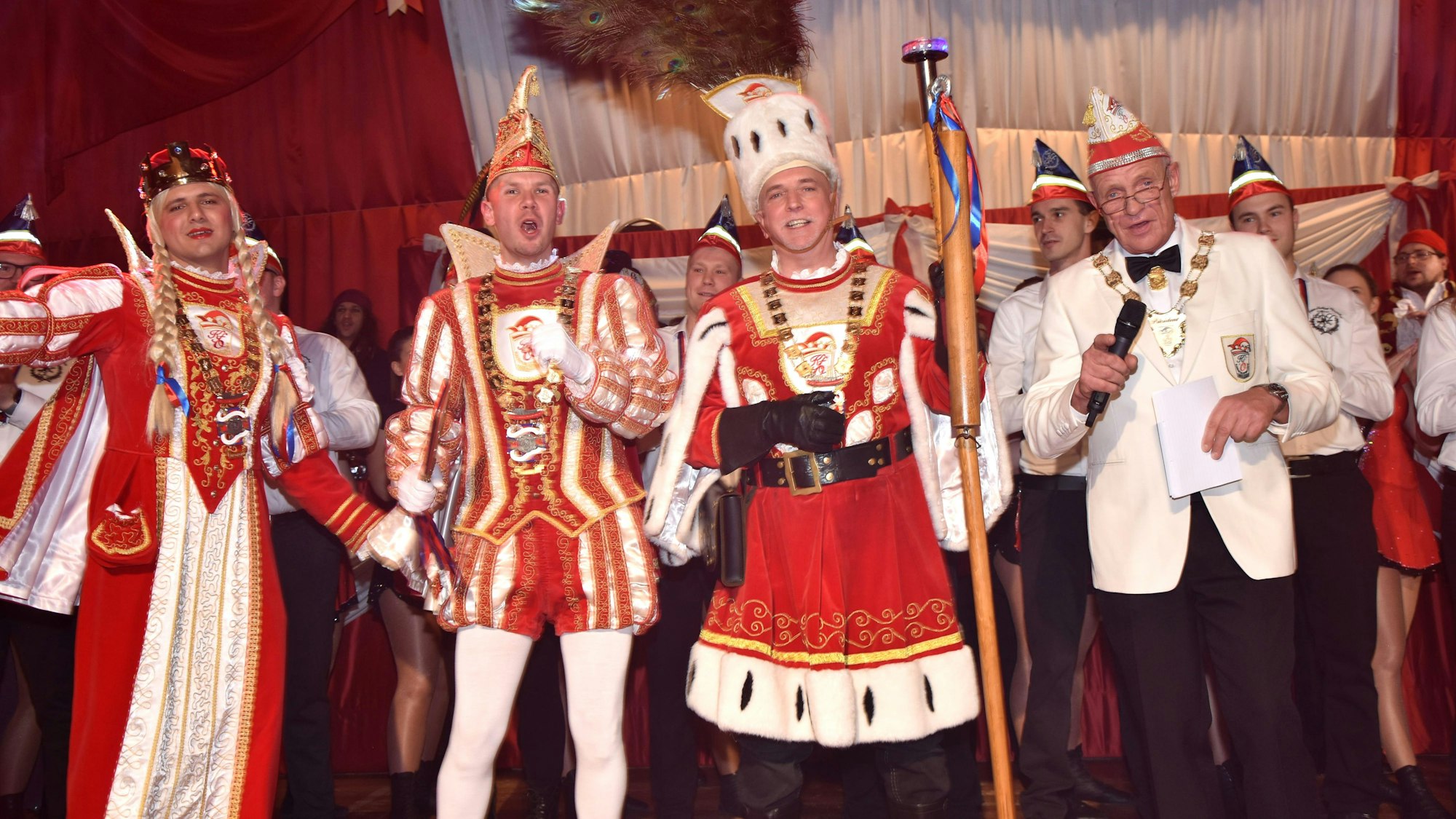 Zu sehen ist ein Dreigestirn auf einer Bühne voller kostümierter Menschen.