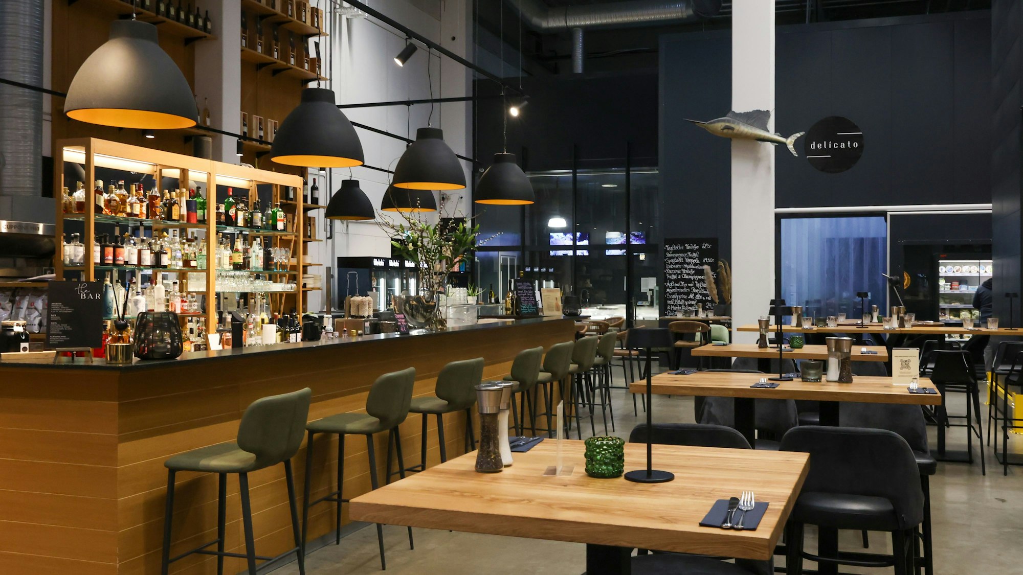 Die Bar mit großen Lampenschirmen darüber ist zu sehen, ebenso einige Hochtische mit Stühlen in dem noch nicht geöffneten Lokal.