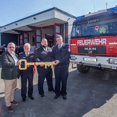 Vier Männer posieren mit einem überdimensionalen Schlüssel vor einem Neubau, rechts im Bild ist ein Einsatzfahrzeug der Feuerwehr zu sehen.