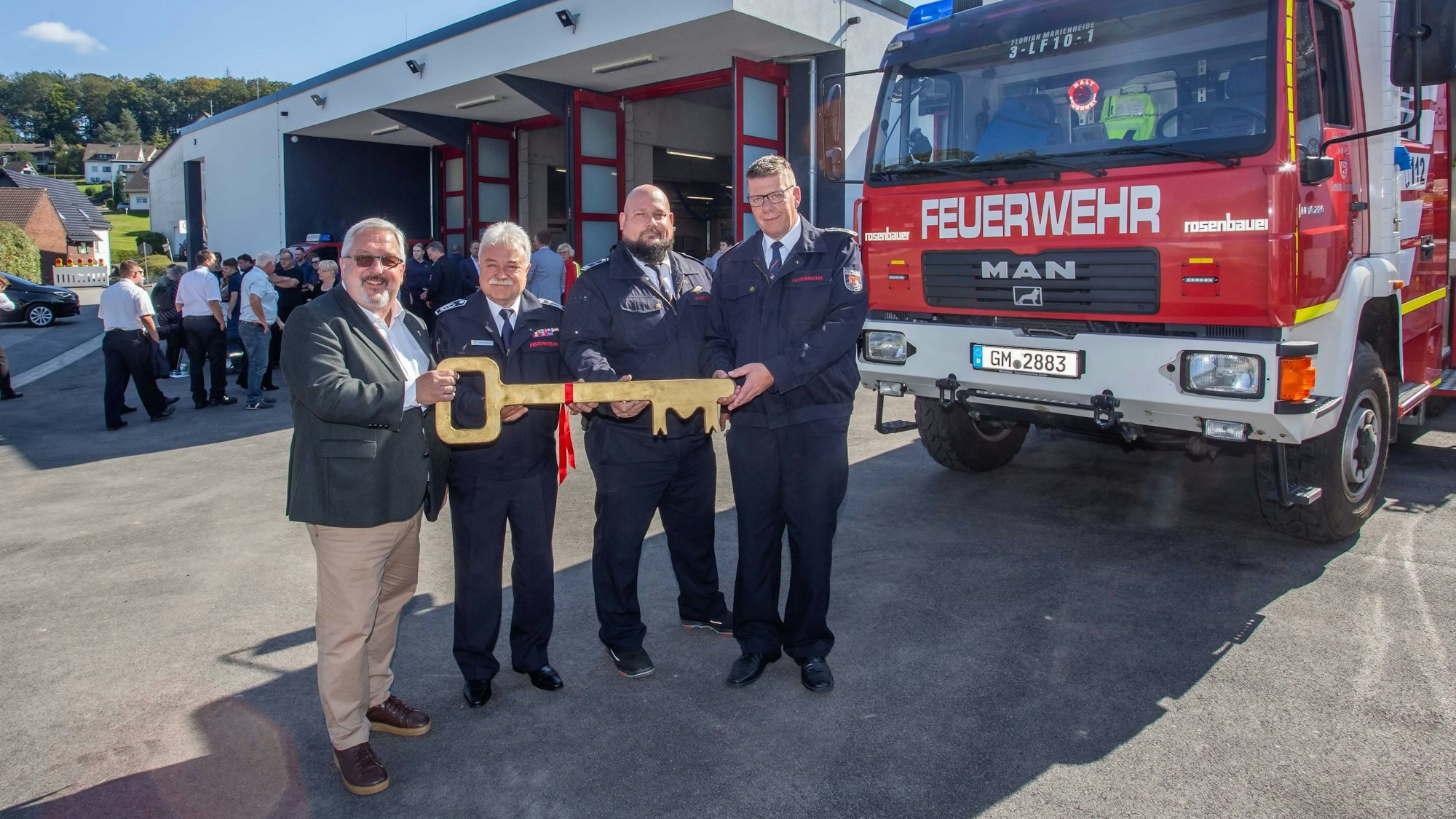 Vier Männer posieren mit einem überdimensionalen Schlüssel vor einem Neubau, rechts im Bild ist ein Einsatzfahrzeug der Feuerwehr zu sehen.