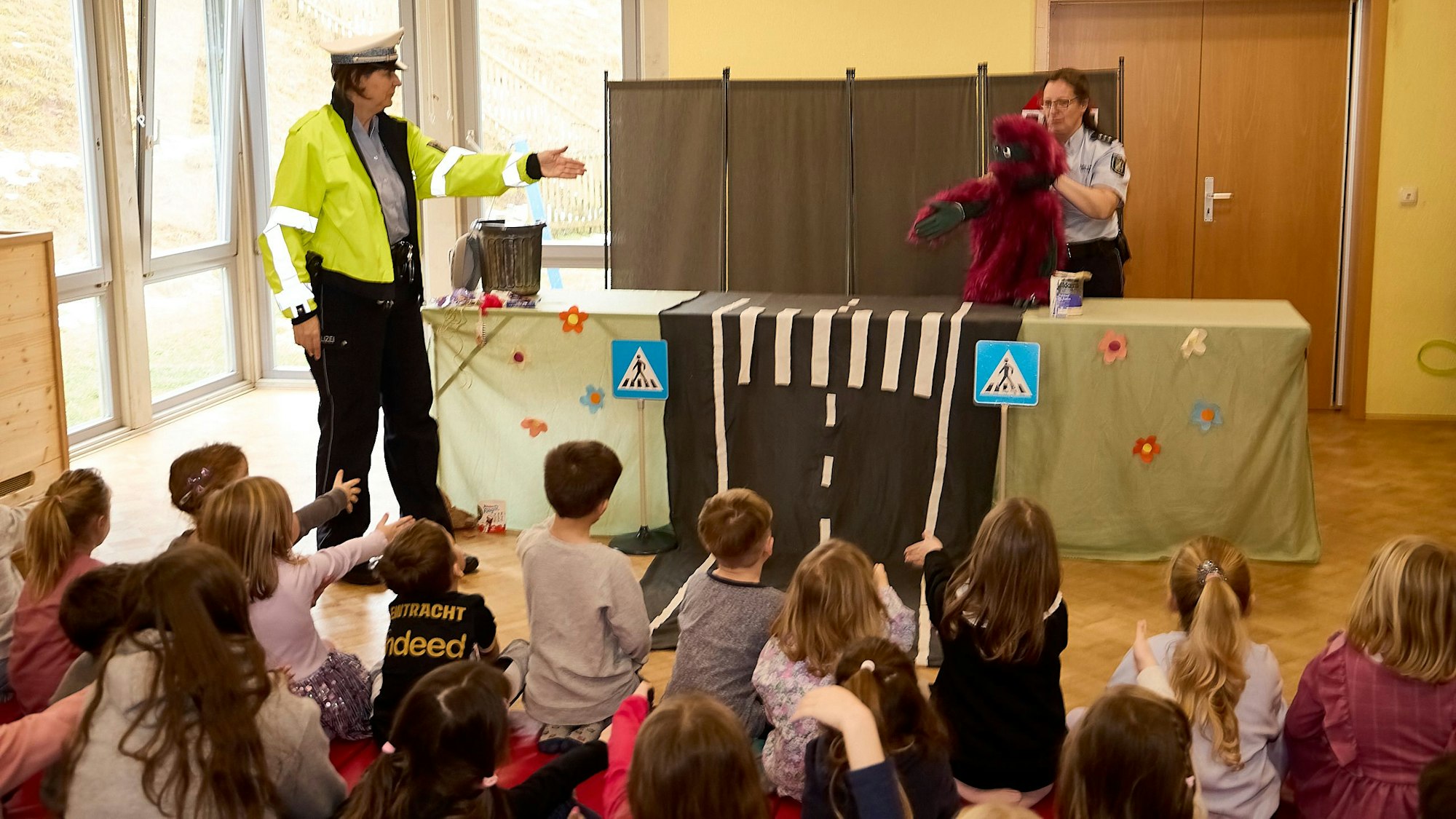 Zahlreiche Kinder sitzen auf dem Boden und schauen in einem Saal einem Theaterstück zu, das zwei Polizistinnen in Uniform mit Handpuppen zeigen.