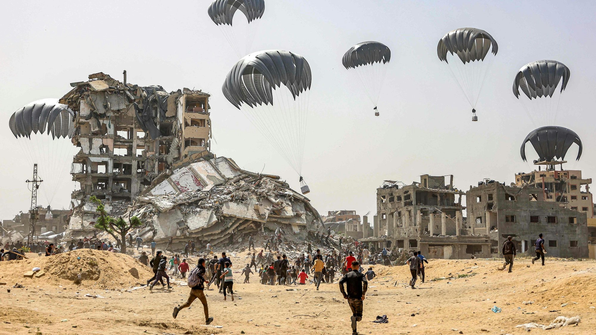 Fallschirme mit Hilfsgütern segeln über ausgebombten Häusern herab. Menschen eilen dorthin, wo sie landen.