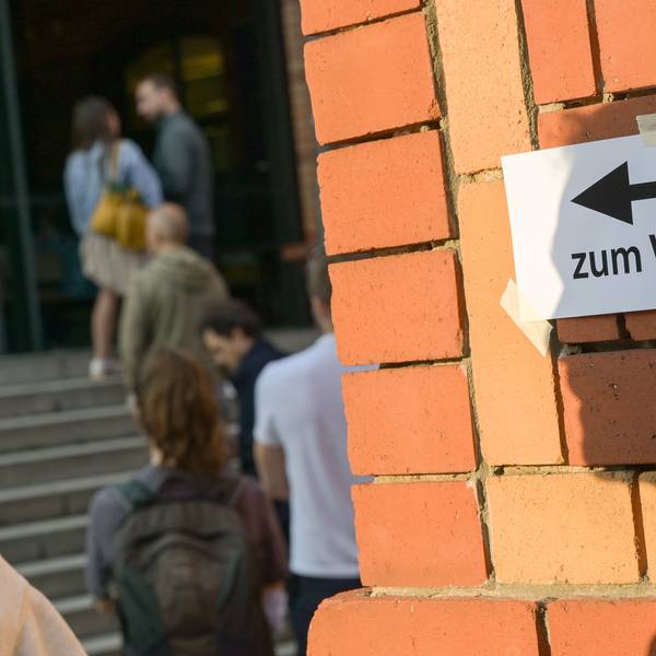 Wählerinnen und Wähler warten vor einem Wahllokal, während auf einer Mauer davor der Schatten eines Mannes auf einen Zettel mit der Aufschrift „Zum Wahllokal“ fällt.
