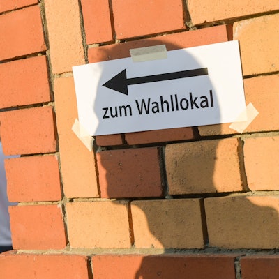 Wählerinnen und Wähler warten vor einem Wahllokal, während auf einer Mauer davor der Schatten eines Mannes auf einen Zettel mit der Aufschrift „Zum Wahllokal“ fällt.