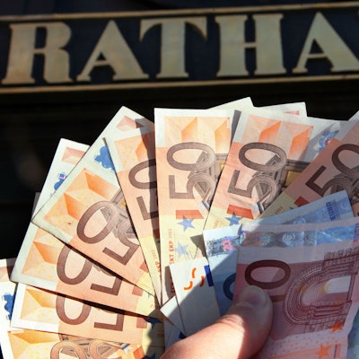 Eine Hand hält Geldscheine, vor allem Fünfzig-Euro-Scheine, vor dem Eingang zu einem Rathaus.