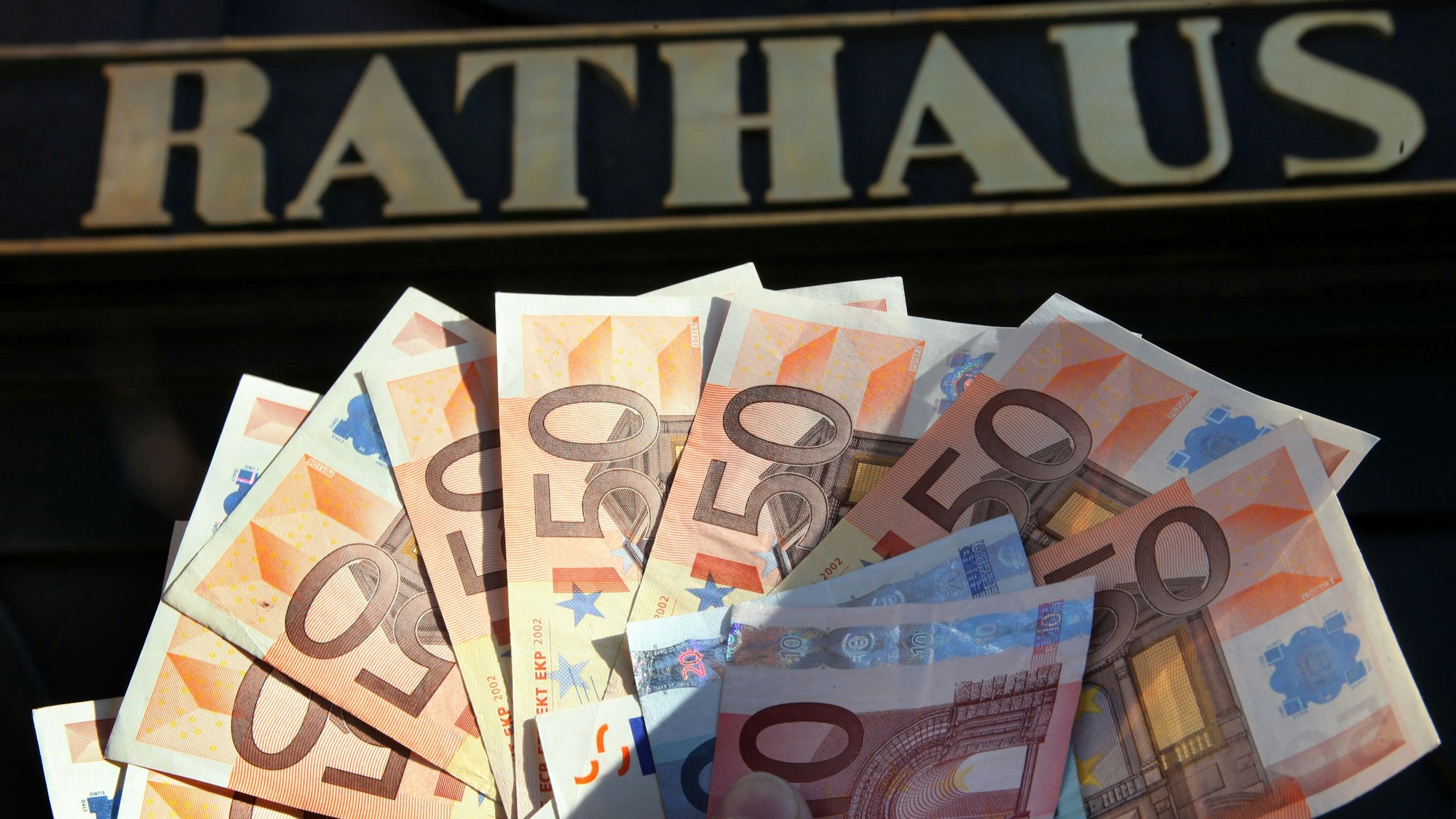 Eine Hand hält Geldscheine, vor allem Fünfzig-Euro-Scheine, vor dem Eingang zu einem Rathaus.