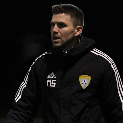 Der Trainer der A-Jugend der JSG Erft 01 Euskirchen, Marcel Schmitz, trägt eine dicke Jacke mit Initialien und Vereinslogo.