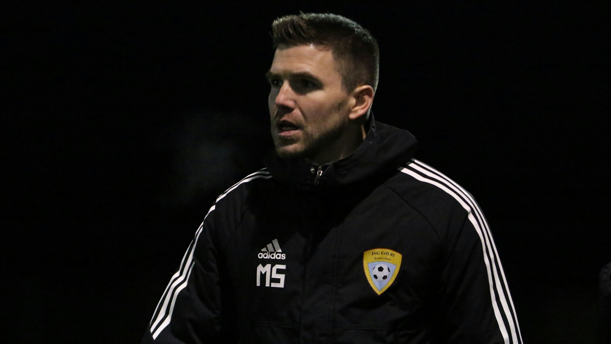 Der Trainer der A-Jugend der JSG Erft 01 Euskirchen, Marcel Schmitz, trägt eine dicke Jacke mit Initialien und Vereinslogo.
