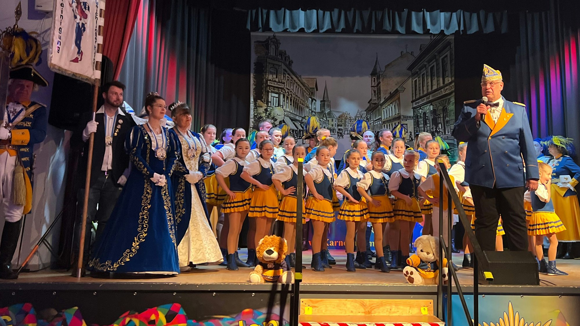 Junge Karnevalisten stehen neben einem Mann am Mikrofon auf einer Bühne.