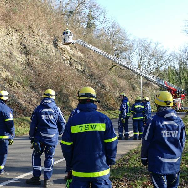 Einsatzkräfte des THW stehen neben einer Drehleiter an einer Straße am Berg.