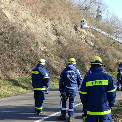 Einsatzkräfte des THW stehen neben einer Drehleiter an einer Straße am Berg.