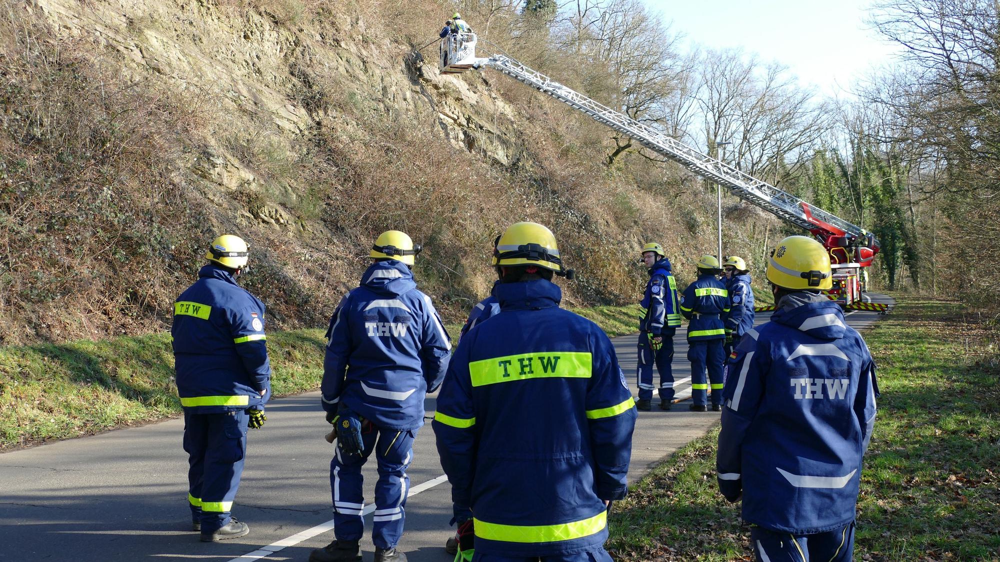 Einsatzkräfte des THW stehen neben einer Drehleiter an einer Straße am Berg.