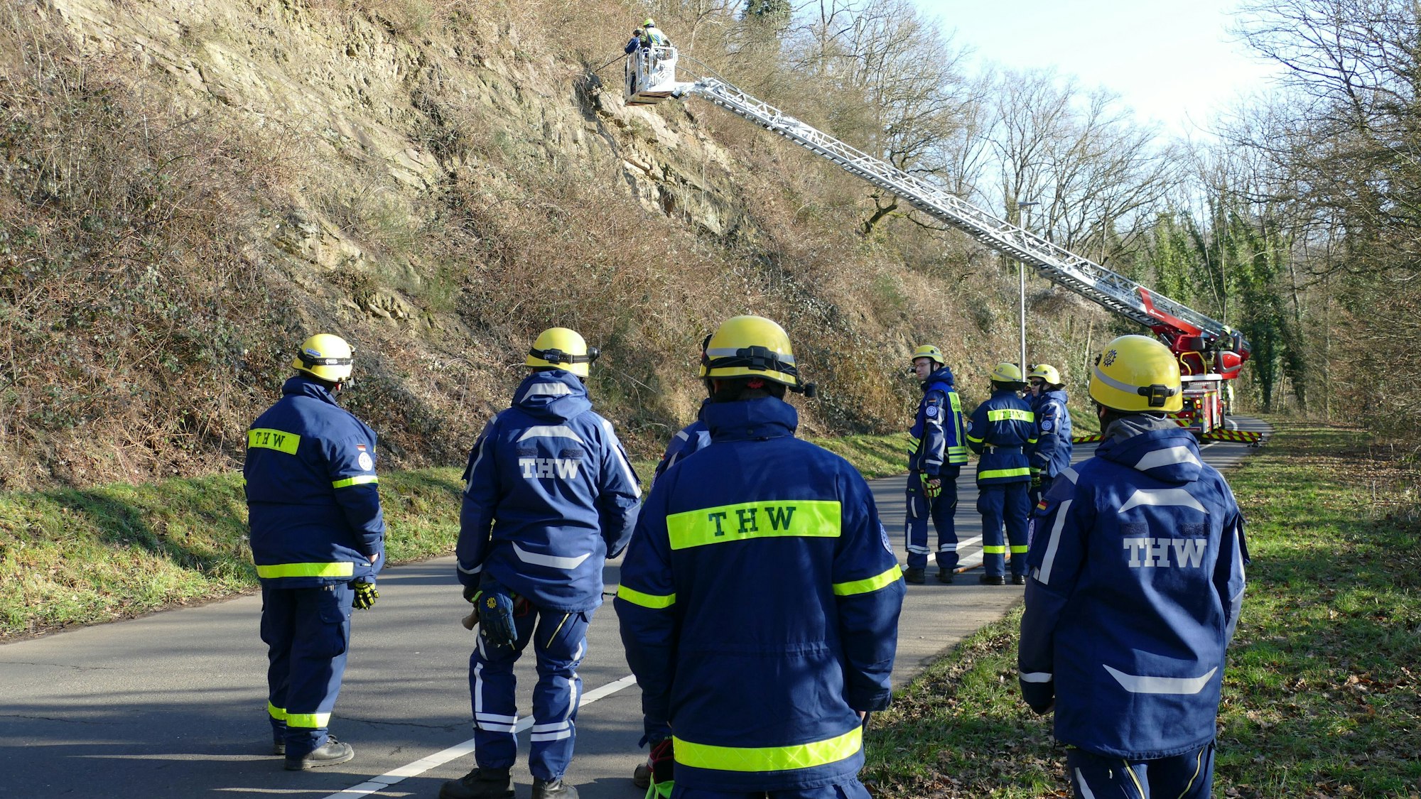 Einsatzkräfte des THW stehen neben einer Drehleiter an einer Straße am Berg.