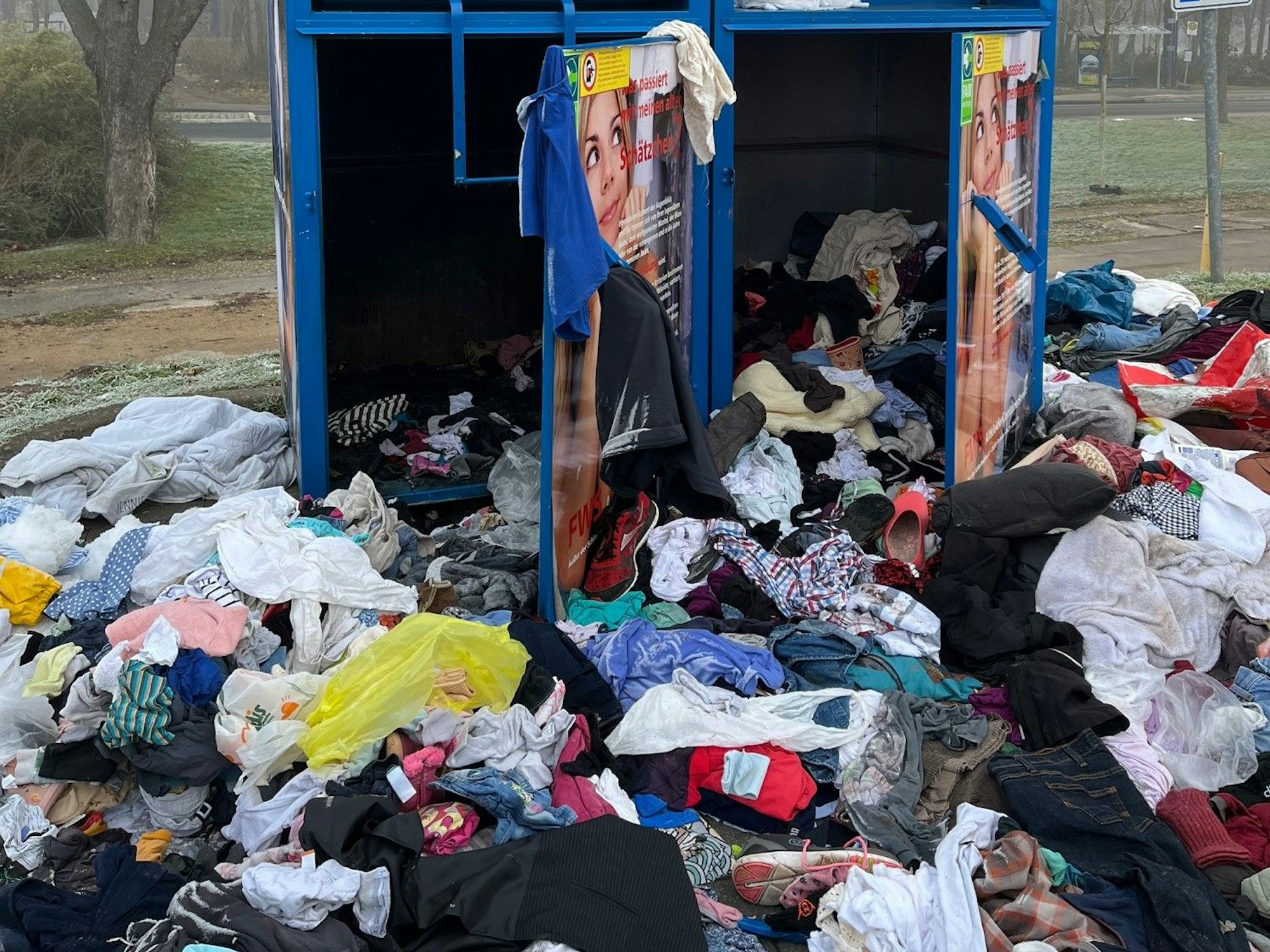 Das Bild zeigt den Altkleider-Container. Die beiden Türen sind aufgebrochen. Vor dem Container liegen zahlreiche Kleidungsstücke.