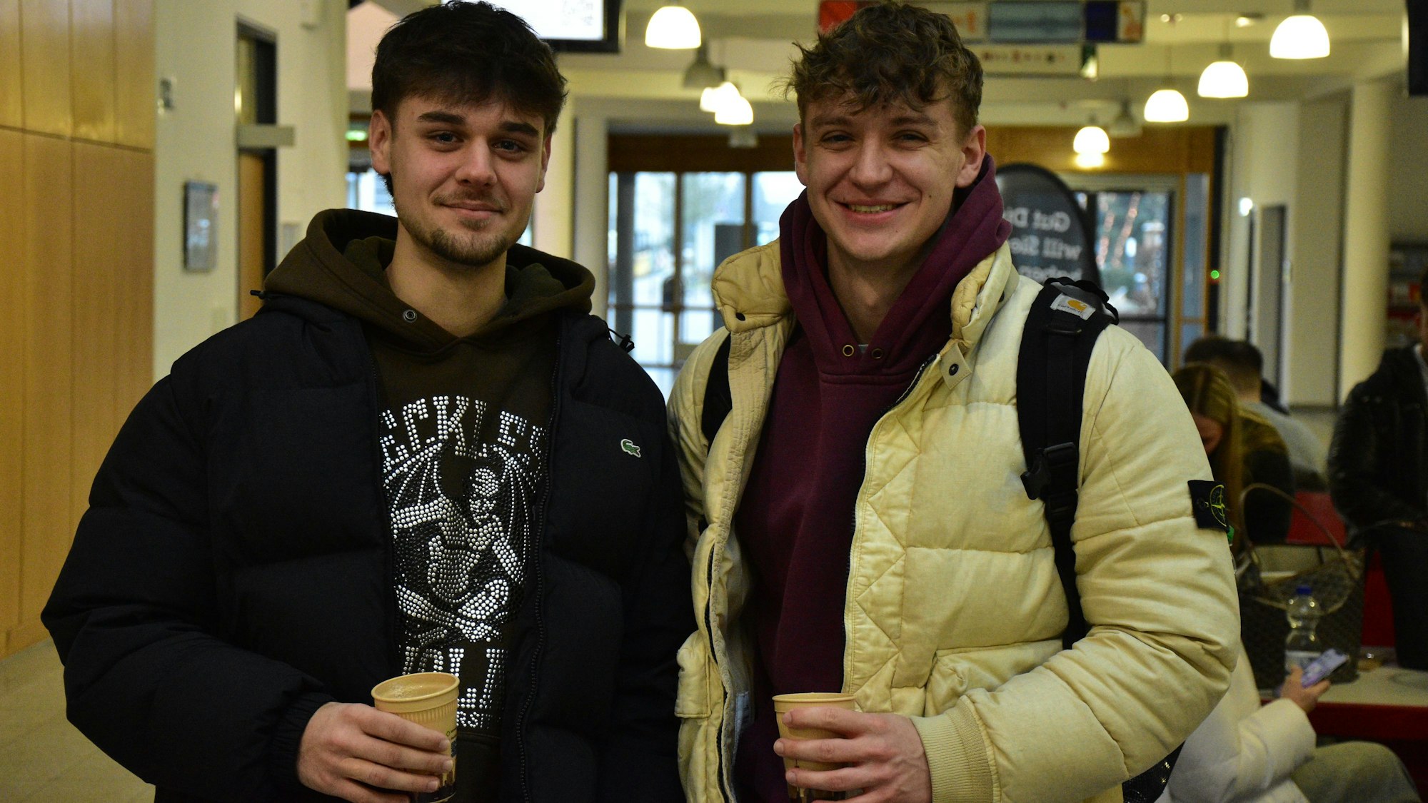 Zwei junge Männer stehen im Flur einer Hochschule, sie haben Kaffeebecher in der Hand.