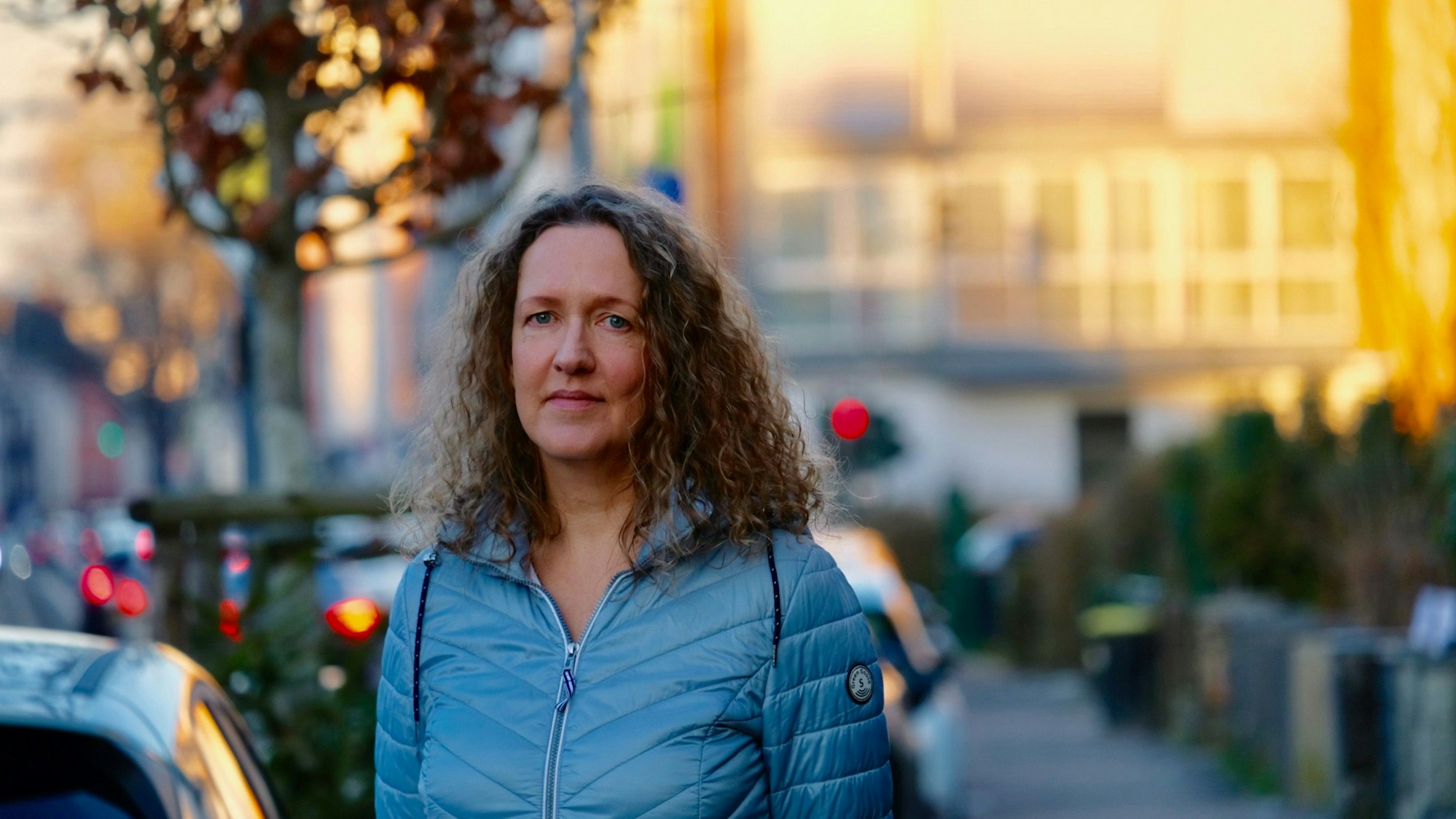 Eine Frau mit langem lockigen Haar steht im Abendlicht auf dem Bürgersteig einer Straße.