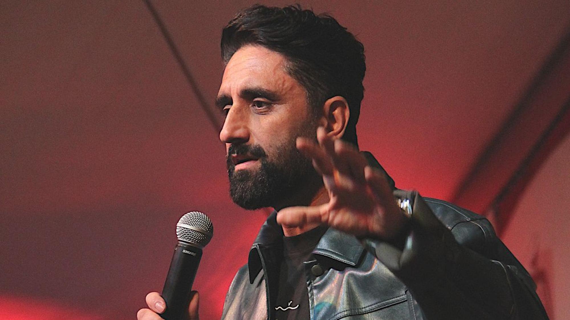 Comedian Djavid trägt eine schwarze Lederjacke und spricht in ein Mikrofon.