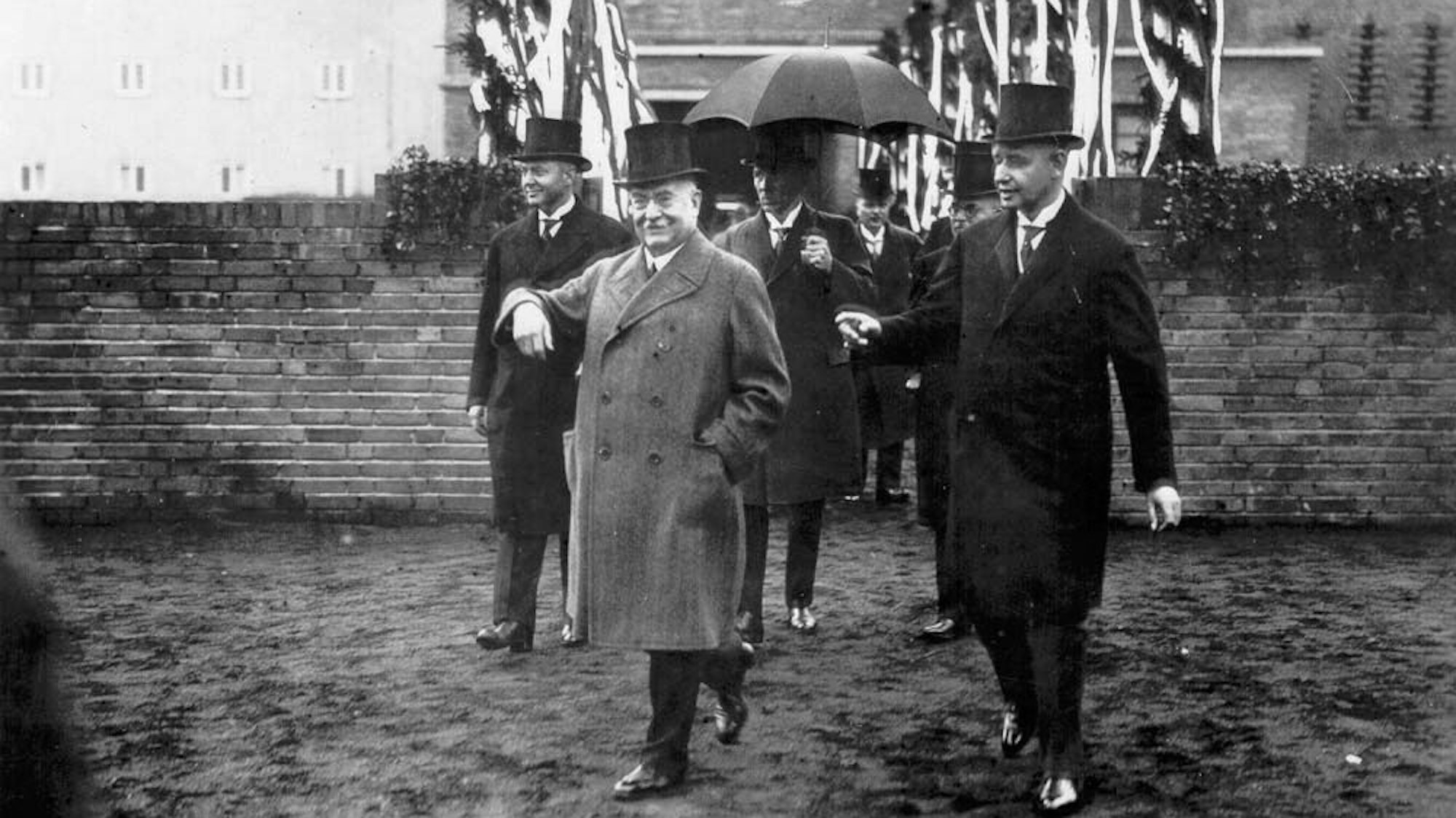 Schwarz-weiß Foto von der Einweihung des CD-Gymnasium: Mehrere Männer laufen vor Backsteinmauer