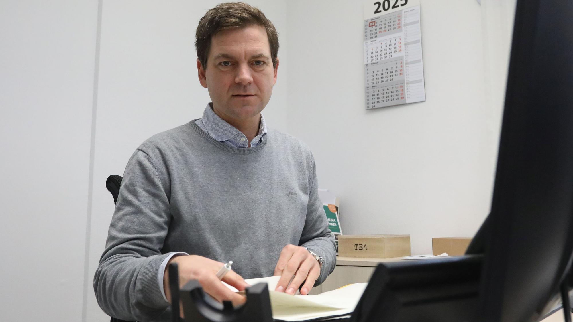 Ein Mann sitzt an einem Schreibtisch vor einem Computermonitor und blättert in einem Stapel Papiere.