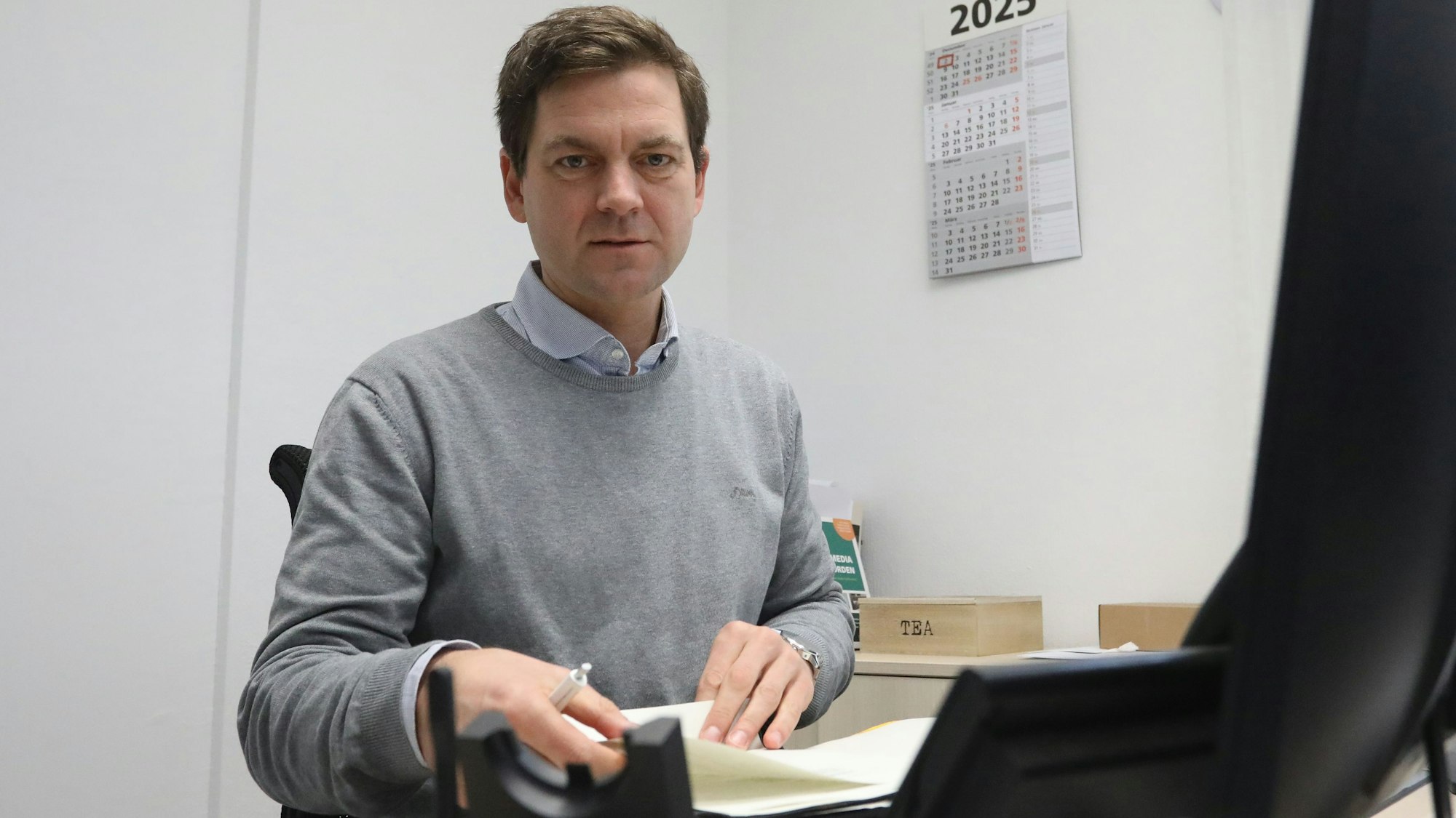 Ein Mann sitzt an einem Schreibtisch vor einem Computermonitor und blättert in einem Stapel Papiere.