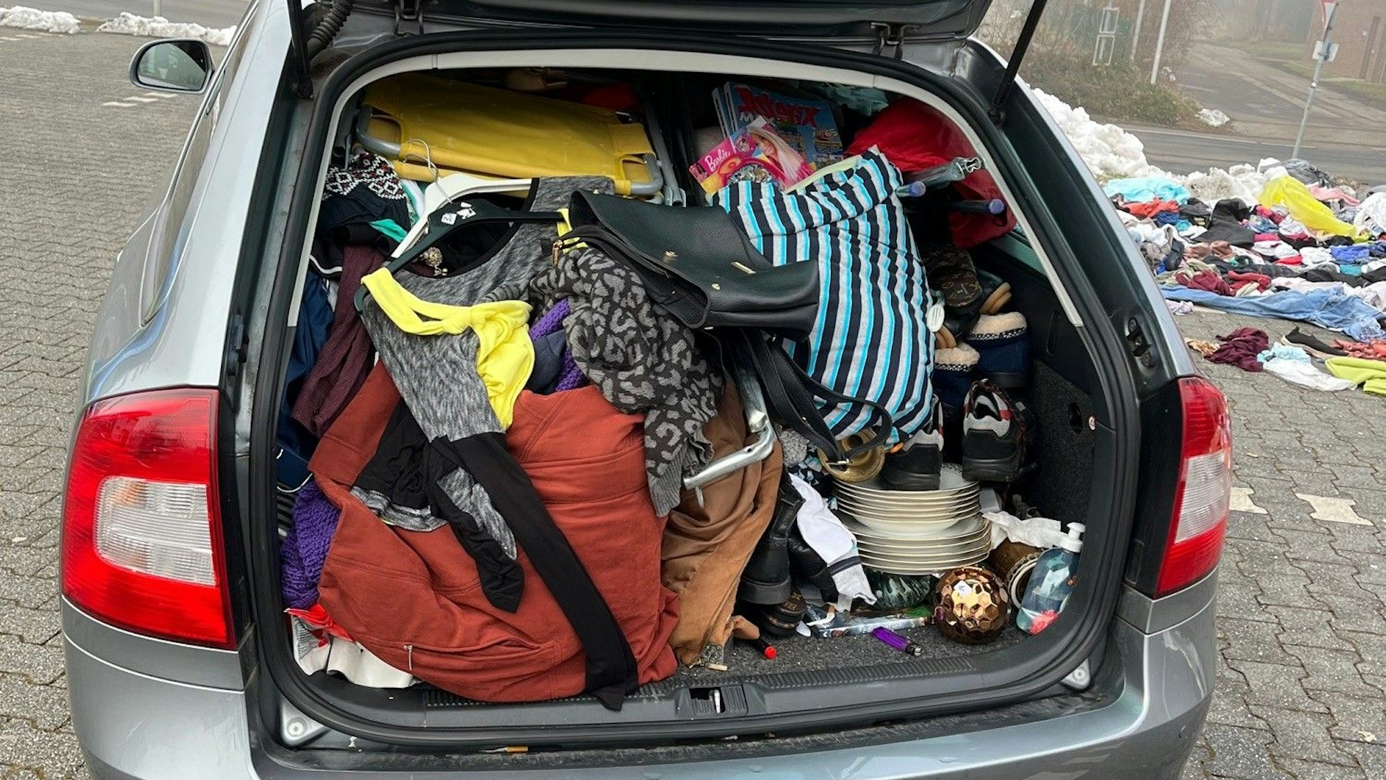 Das Bild zeigt den Kofferraum eines Autos. Die Heckklappe ist offen. In dem Kofferraum sind zahlreiche Kleidungsstücke gestapelt.