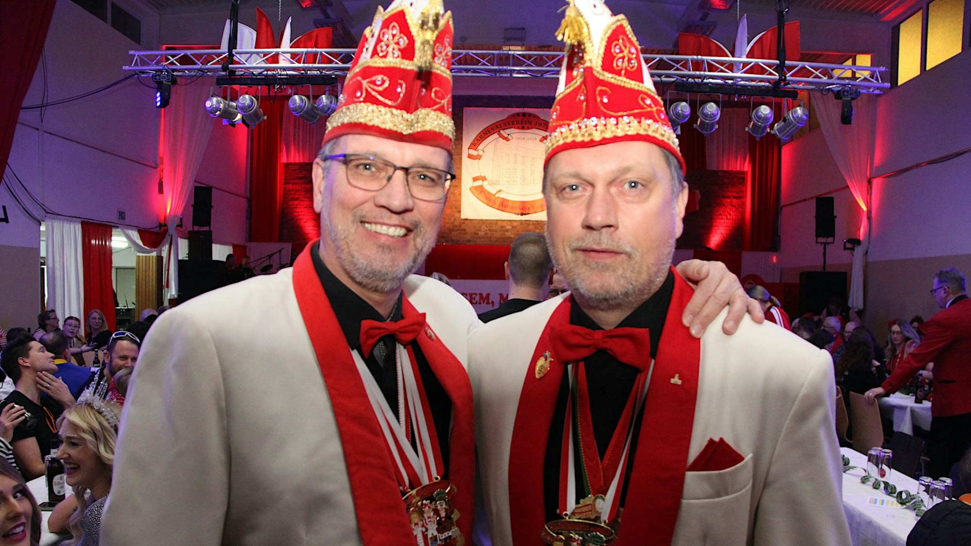 Zwei Männer in Narrenkappen und weiß-roten Jacketts stehen Arm in Arm in einem Saal.