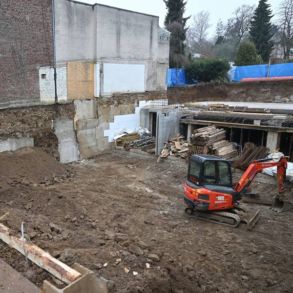 Blick von oben in die Baugrube. Man sieht einen Erdhaufen vor der Hauswand, Baumaterial sowie einen Bagger.