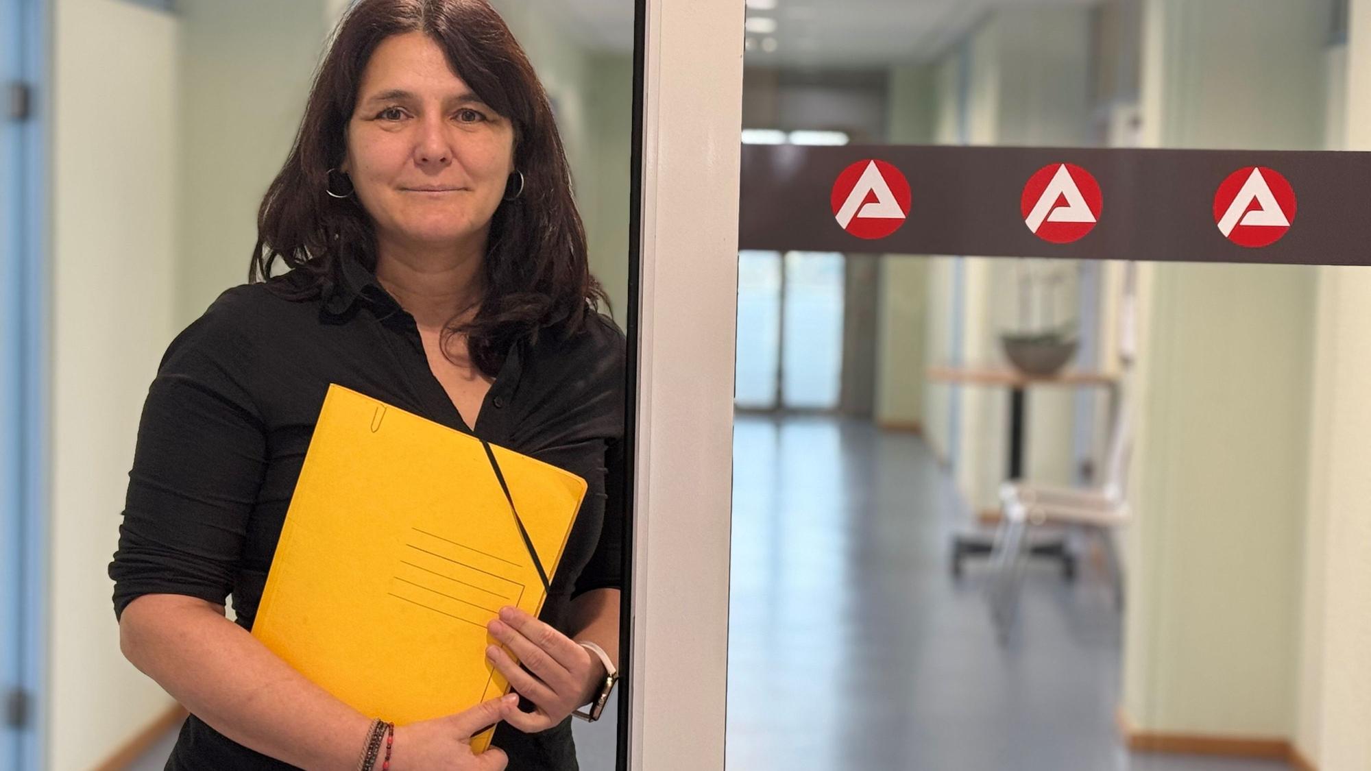 Anja Daub ist Geschäftsführerin operativ der Agentur für Arbeit in Brühl. Sie hält eine gelbe Aktenmappe in den Händen und steht an einer Glastür mit drei Logos der Arbeitsagentur.