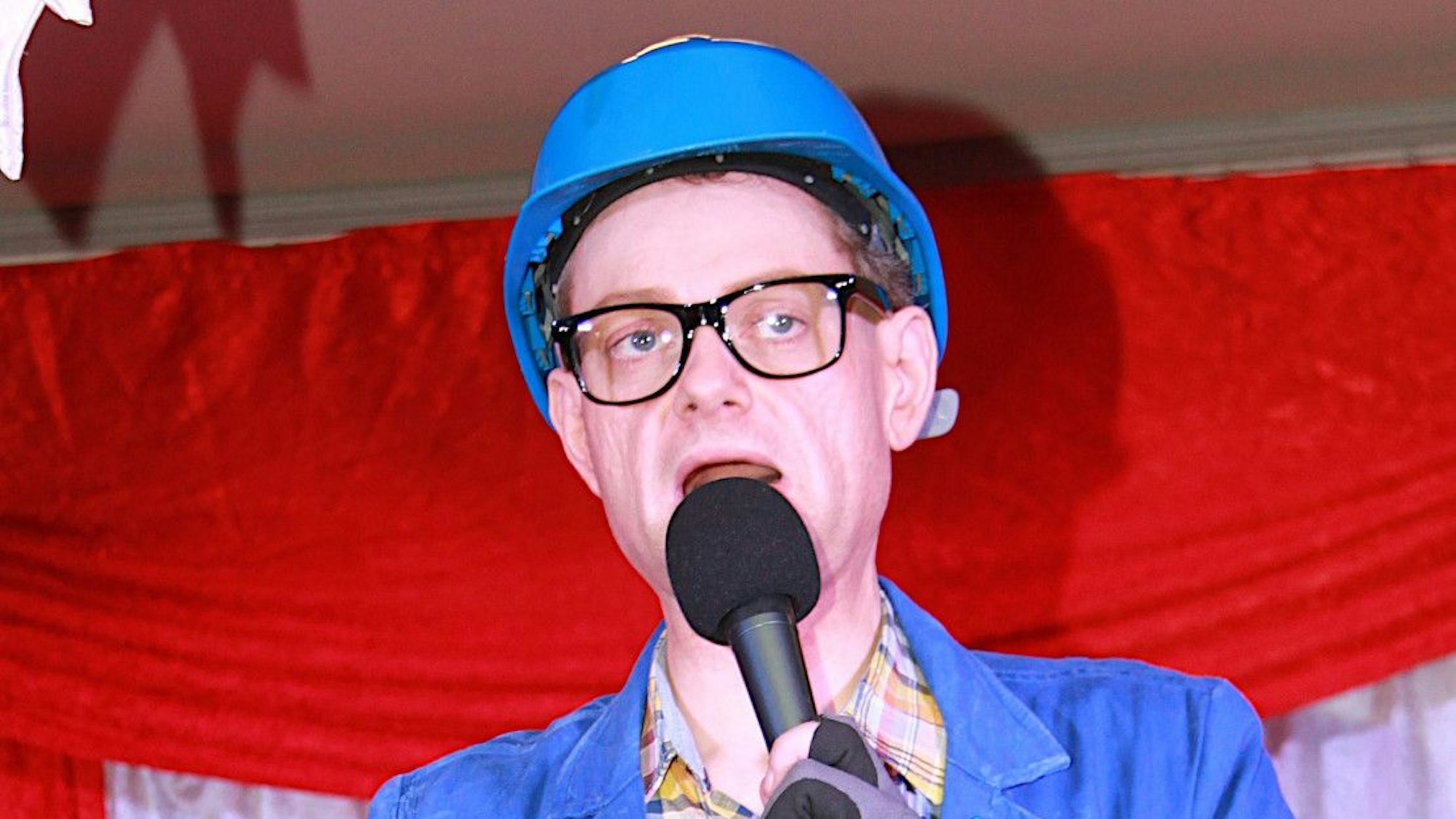 Der Büttenredner trägt einen blauen Schutzhelm, ein kariertes Hemd und eine blaue Jacke.