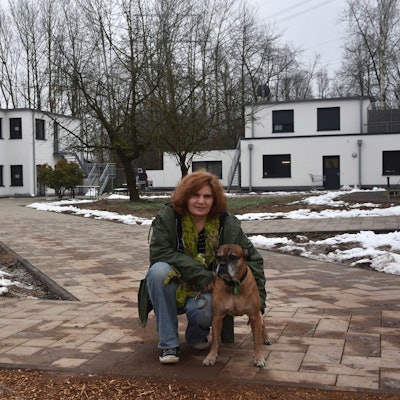 Zu sehen ist eine Frau mit einem alten Boxerhund vor neuen Gebäuden.