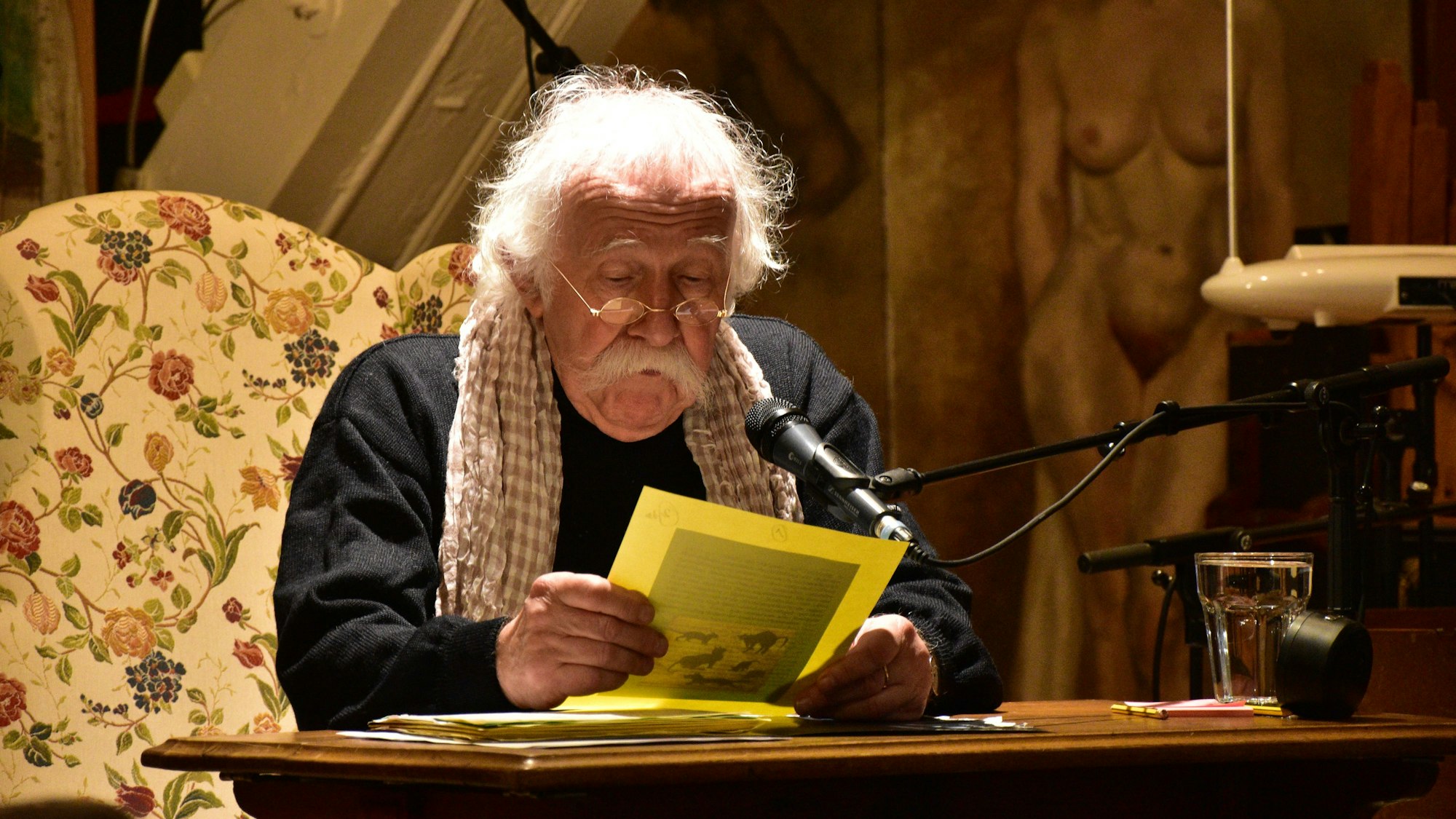 Ein Mann mit weißen Haaren und einem breiten weißen Schnurrbart rezitiert an einem Tisch.