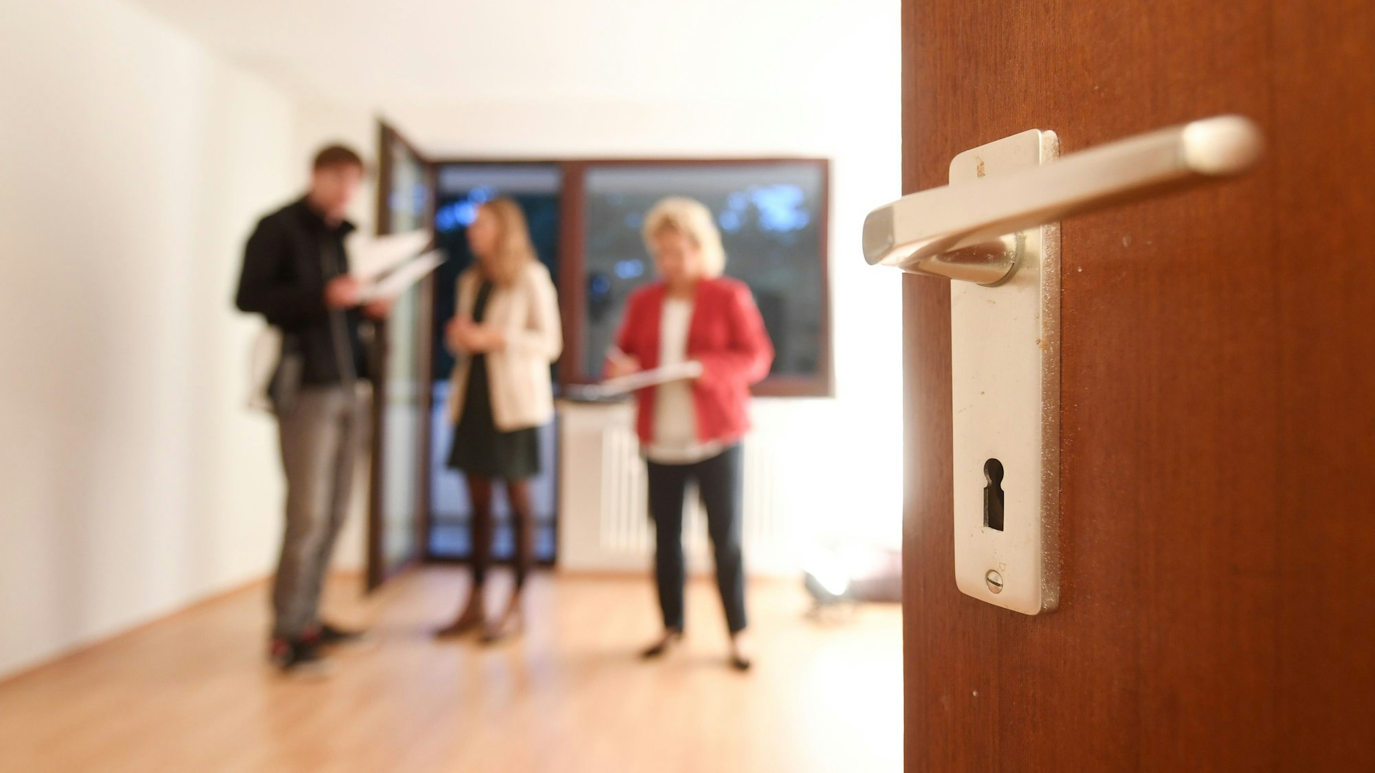 Eine Maklerin spricht mit Interessenten bei einer Wohnungsbesichtigung, während im Vordergrund eine Türklinke zu sehen ist.