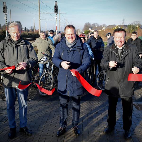Das Foto zeigt drei Männer mit Scheren, die ein rotes Band durchgeschnitten haben, vor einer Gruppe von Radfahrern.