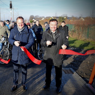 Das Foto zeigt drei Männer mit Scheren, die ein rotes Band durchgeschnitten haben, vor einer Gruppe von Radfahrern.