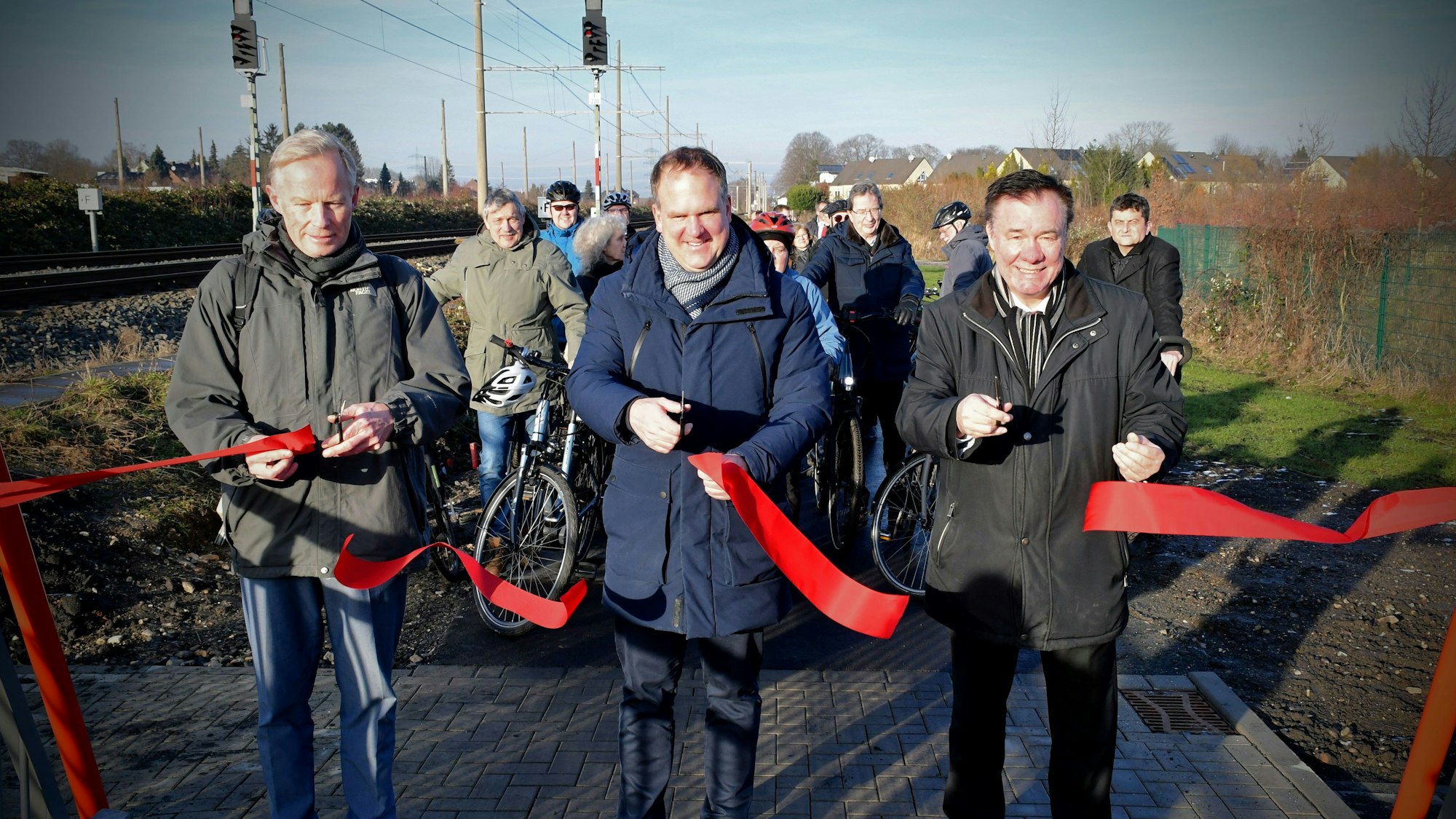 Das Foto zeigt drei Männer mit Scheren, die ein rotes Band durchgeschnitten haben, vor einer Gruppe von Radfahrern.