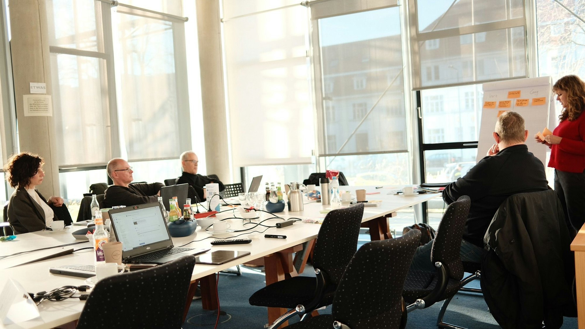 Menschen sitzen an einem Tisch, Computer stehen vor ihnen, eine Frau steht an einer Flipchart.