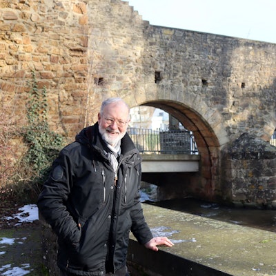 Norbert Liebing steht außerhalb der Stadtmauer und legt die Hand auf die Erftmauer. Im Hintergrund sind die Rundbögen der Schoßpforte zu erkennen.