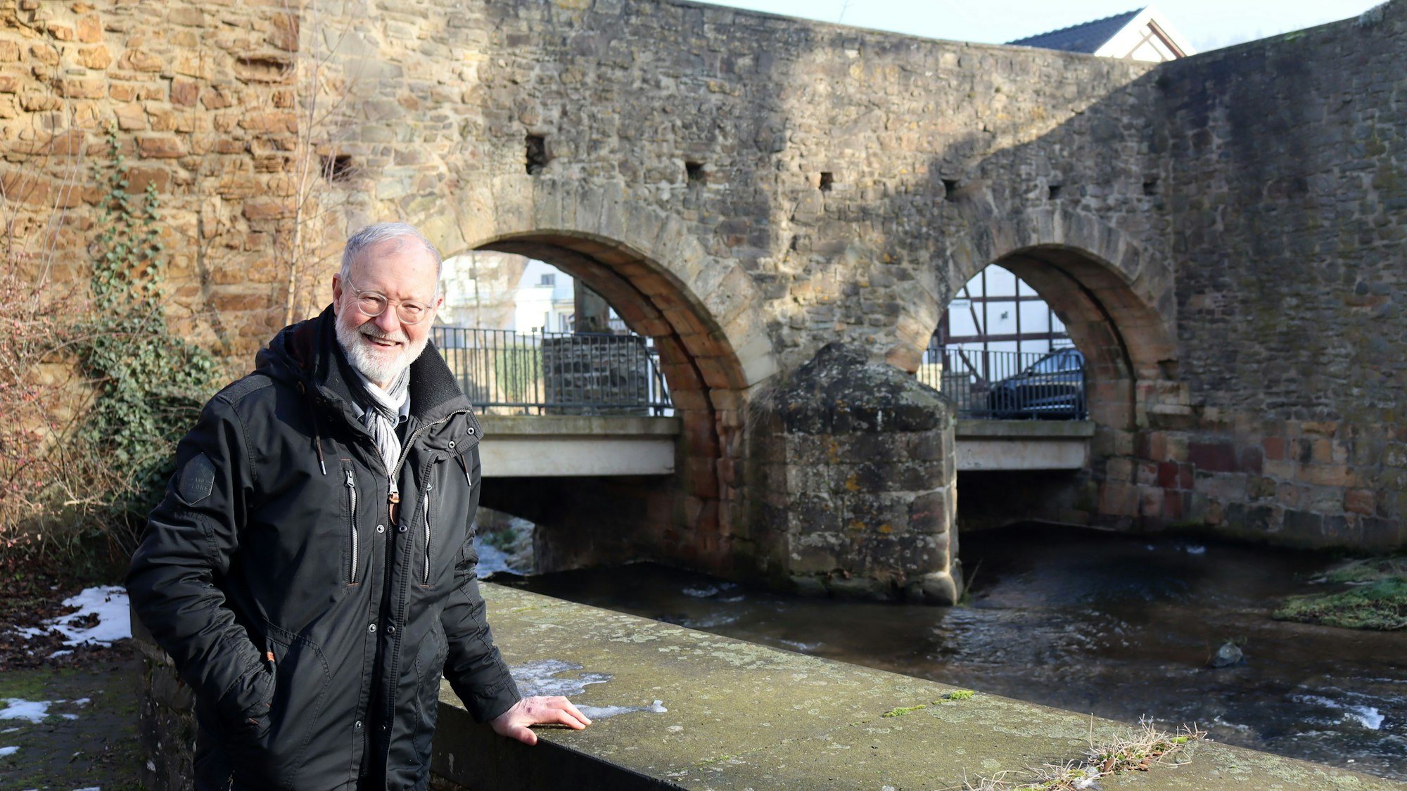 Norbert Liebing steht außerhalb der Stadtmauer und legt die Hand auf die Erftmauer. Im Hintergrund sind die Rundbögen der Schoßpforte zu erkennen.