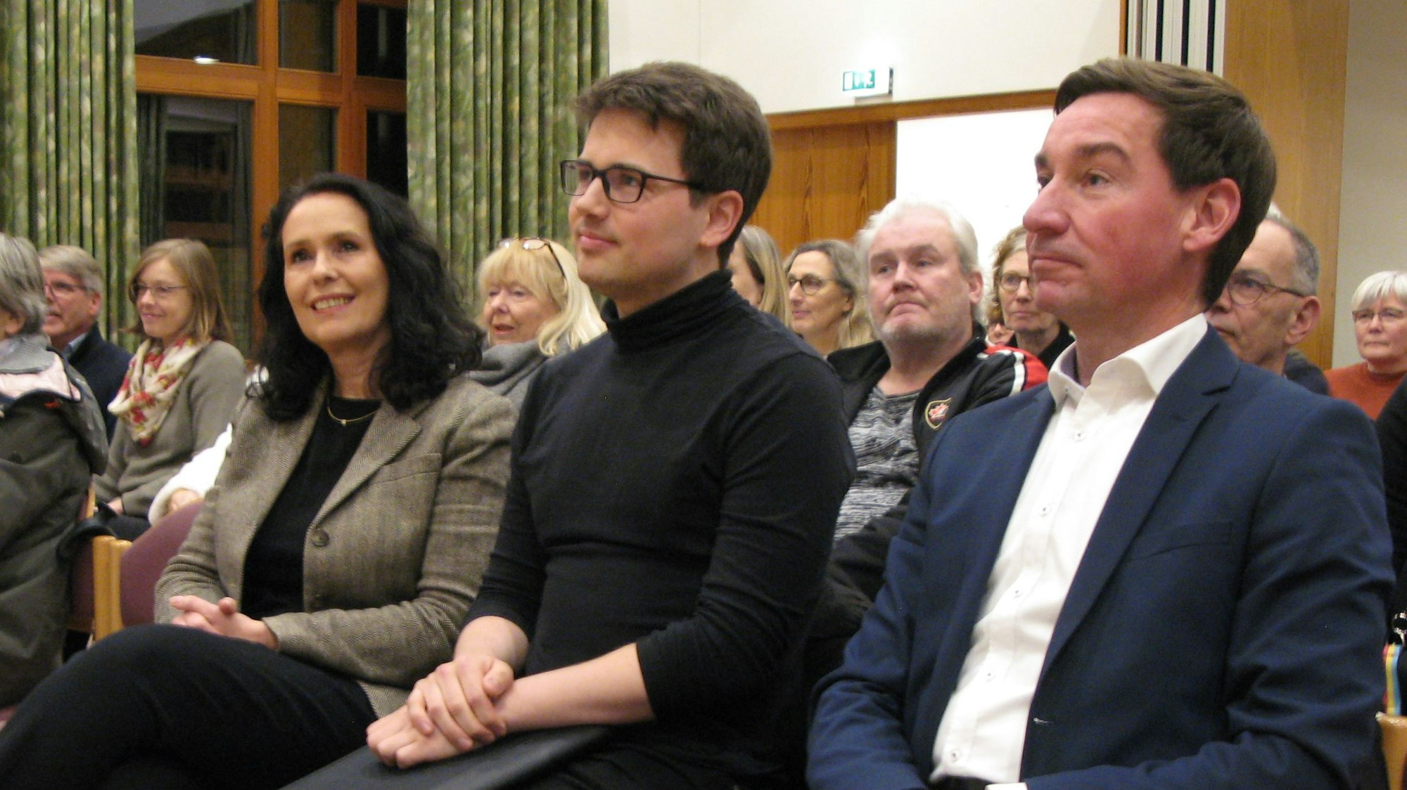 Zwei Männer und eine Frau sitzen in der erste Reihe in einem Saal.