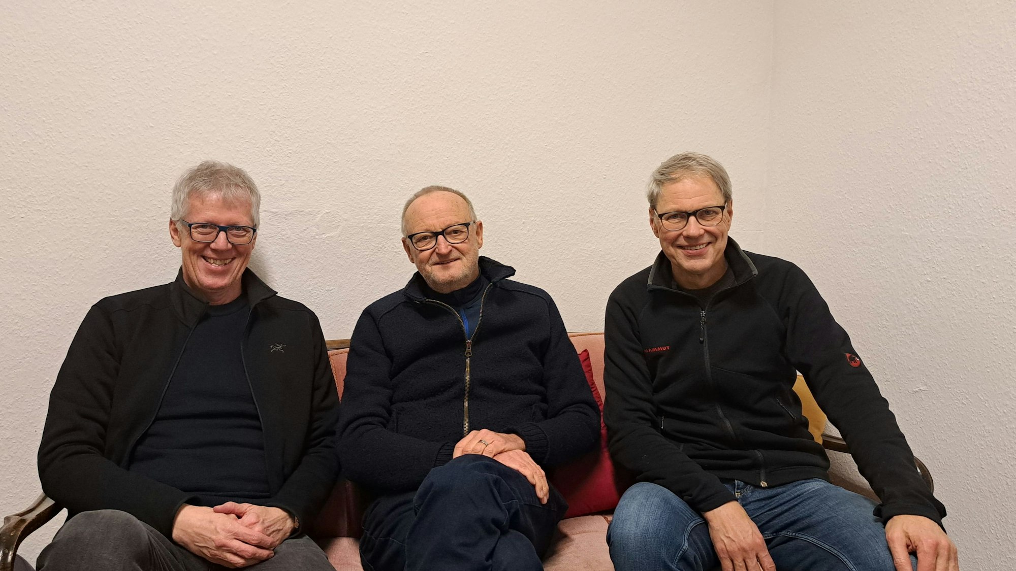 Drei Männer, die Brillen und schwarze Jacken tragen, sitzen auf einer Couch und lachen in die Kamera