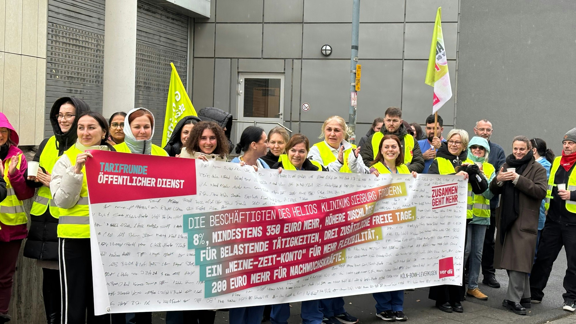Pflegekräfte stehen vor dem Helios Krankenhaus in Siegburg und halten ein Banner. Darauf steht „Tarifrunde Öffentlicher Dienst“.