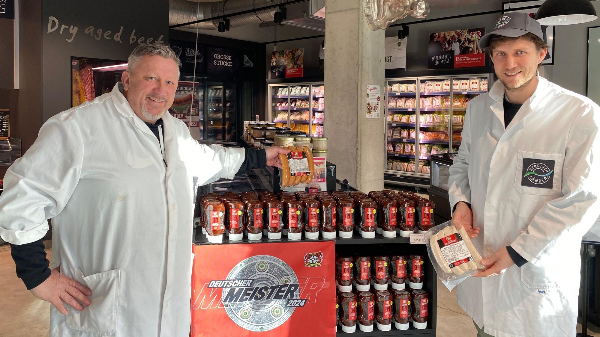 Uwe Nickut und Marcel Schröder stehen in weißen Kittel mit Stadionwurst und Ktakauer in der Hand vor einem Currysoßenstand in dem Markt „Bergischländer“.